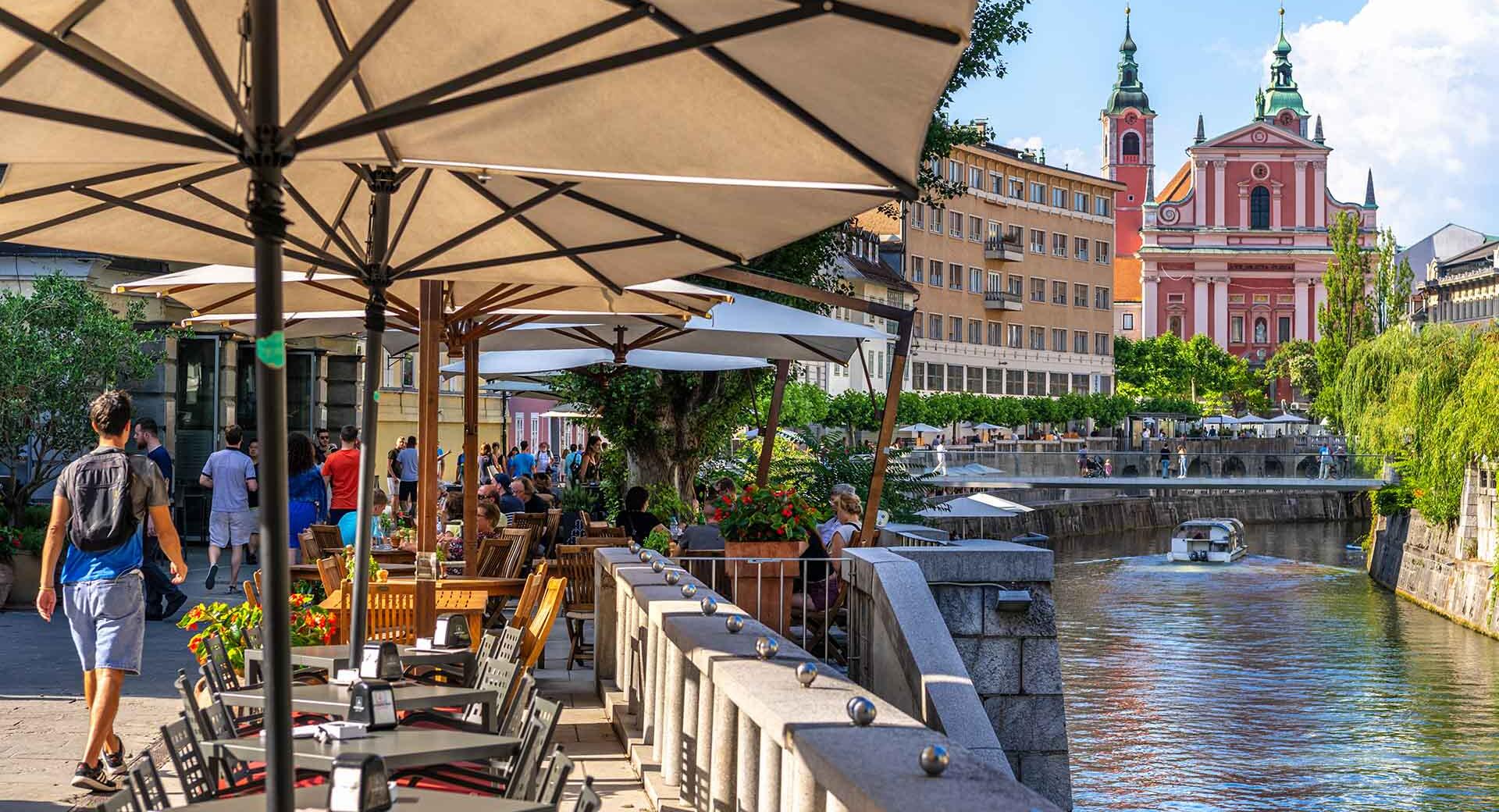 Tables, umbrellas, and chairs on the riverbank. In the background, a bridge and a church.