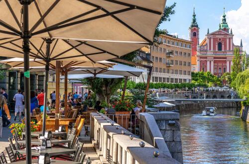 Tables, umbrellas, and chairs on the riverbank. In the background, a bridge and a church.