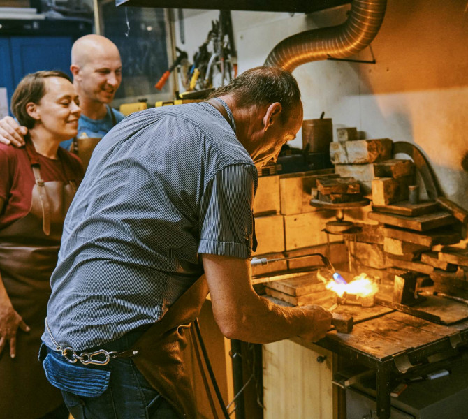 Ein Goldschmied schmilzt in der Werkstatt mit einem Gasbrenner Metall, waehrend zwei Teilnehmende den Herstellungsprozess von Schmuck beobachten.