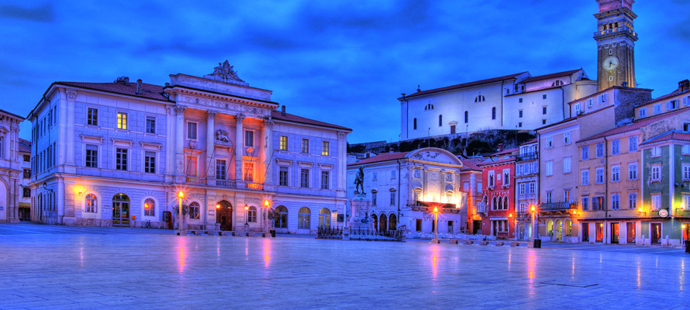 Prostoren kamnit trg, obdan z zgodovinsko arhitekturo, ob mraku.