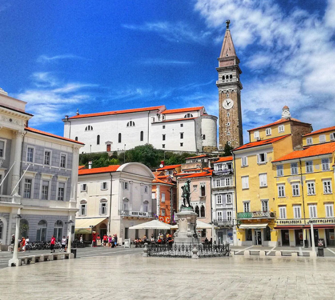 Der Tartini-Platz in Piran im Sommer mit der Statue in der Mitte, bunten Häusern im Hintergrund und einem hohen Kirchturm unter blauem Himmel.