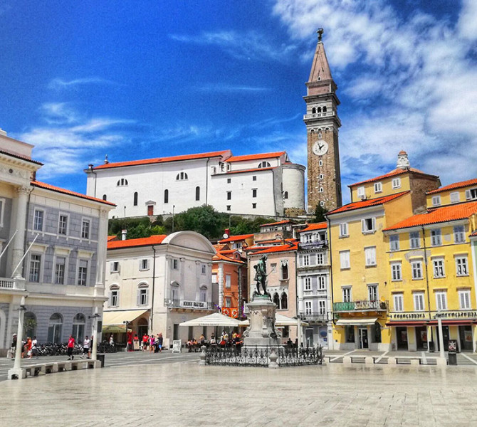 Pogled na Tartinijev trg v Piranu s spomenikom Giuseppeja Tartinija, beneškimi stavbami, zvonikom in cerkvijo sv. Jurija v ozadju pod modrim nebom.