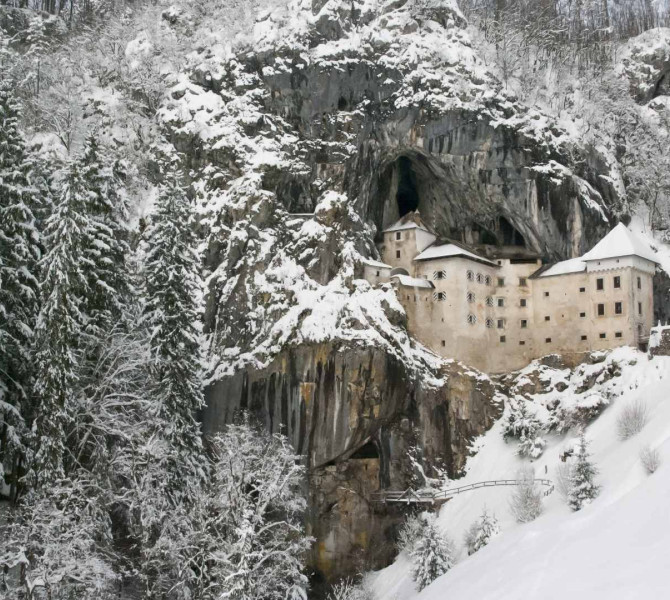 Predjamski grad v zimski pokrajini pod visoko skalno steno, obdan z zasnezenim gozdom.