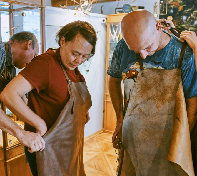 Zwei Mentoren in einer Goldschmiedewerkstatt helfen zwei Teilnehmenden, vor Arbeitsbeginn lederne Schutzschuerzen anzulegen.