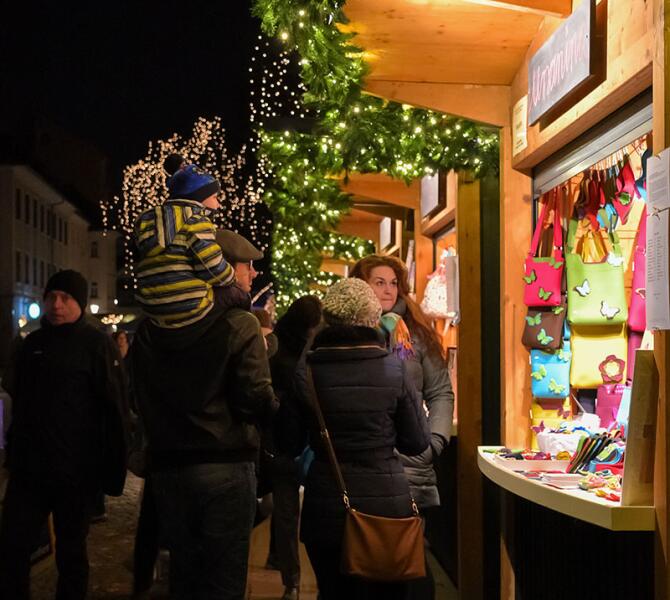 People enjoying a festive market, with illuminated stalls and decorative lights, browsing colourful items on display.