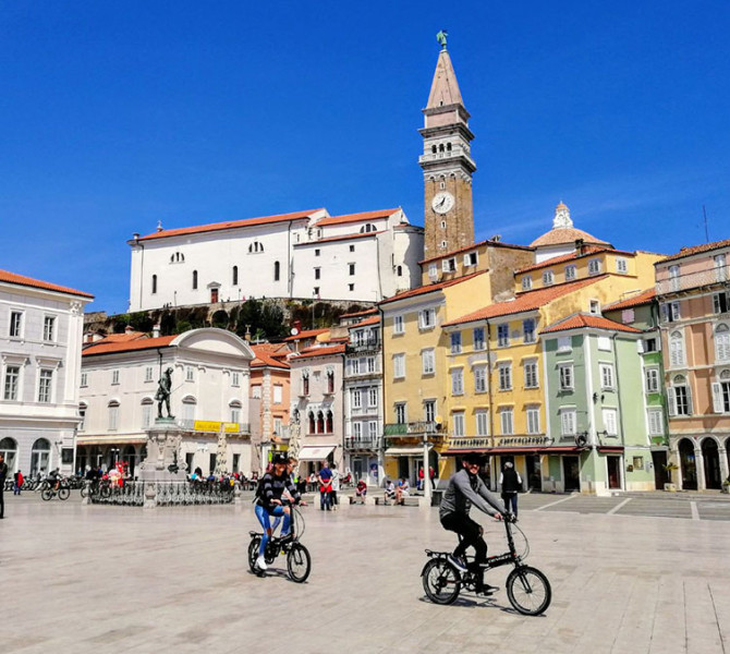 Two cyclists in Tartini Square in Piran, with the Town Hall and other buildings in the background.