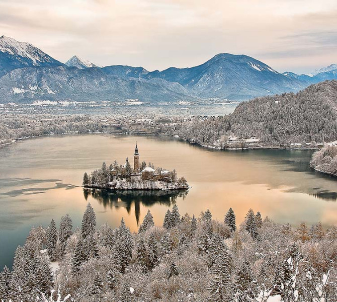 Panoramska slika Blejskega jezera in otoka pozimi. V ozadju zasnežene gore.