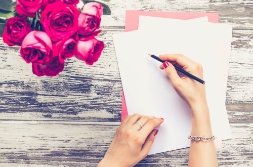  Hands of a woman, a pencil in the right, the left one on a sheet of a paprer, a bouquet of red roses next to it.