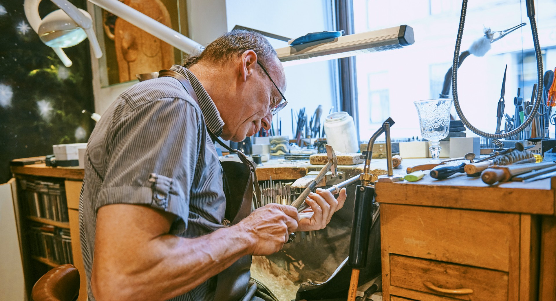 Starejsi zlatar sedi za delovno mizo in obdeluje kovino z rocnim orodjem.