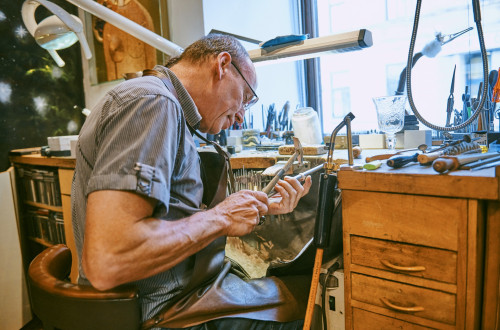 Starejsi zlatar sedi za delovno mizo in obdeluje kovino z rocnim orodjem.