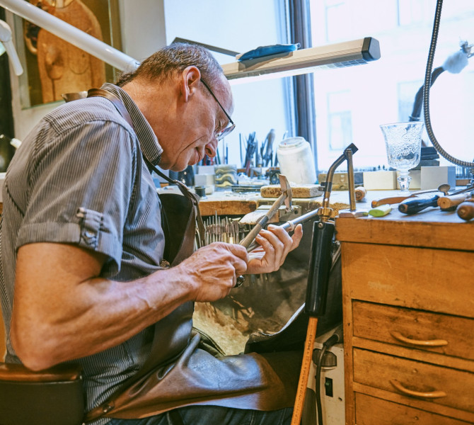 Un anziano orafo è seduto al banco da lavoro e lavora il metallo con uno strumento manuale.