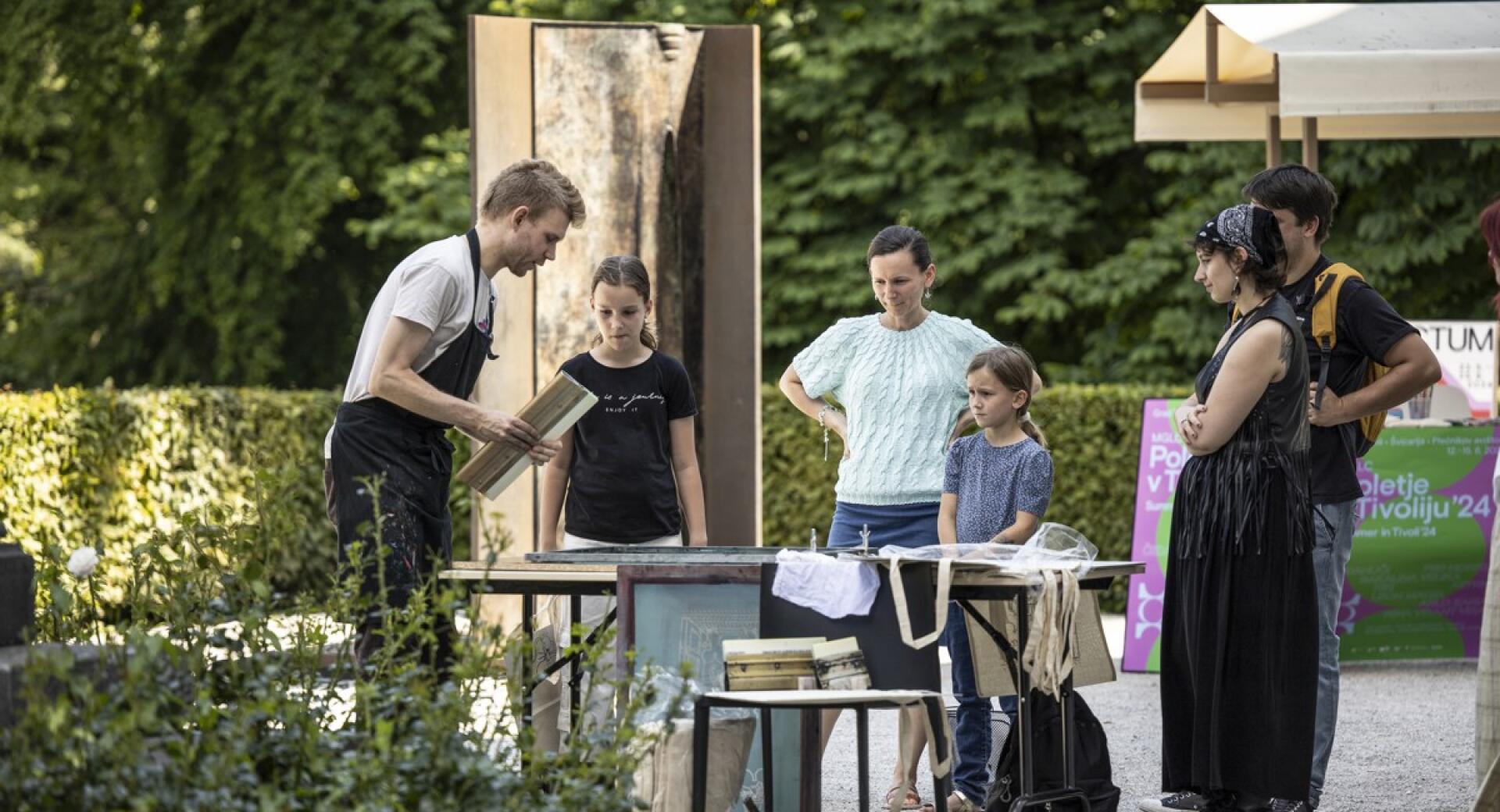 A facilitator demonstrates a creative process at an outdoor worktable to a small group of visitors, including children.