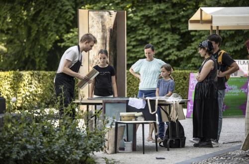 A facilitator demonstrates a creative process at an outdoor worktable to a small group of visitors, including children.