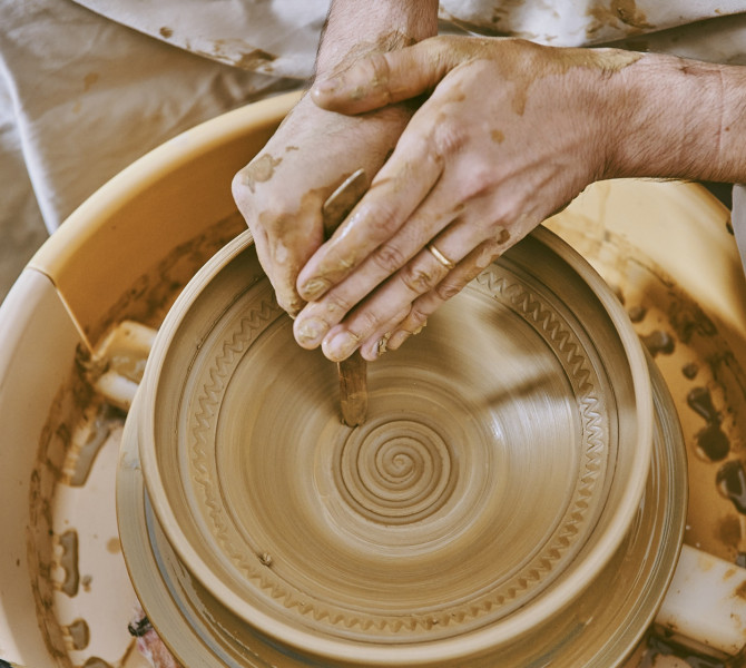 Les mains d’un participant à un atelier de poterie façonnent l’argile sur un tour de potier en rotation et tracent un motif en spirale avec un outil.