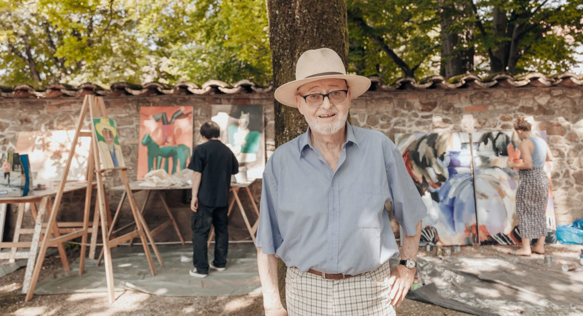 Moski s slamnikom stoji pred drevesom, za drevesom pa so platna in slikarji.