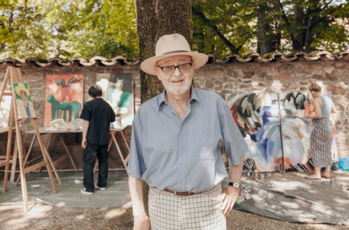 Moski s slamnikom stoji pred drevesom, za drevesom pa so platna in slikarji.