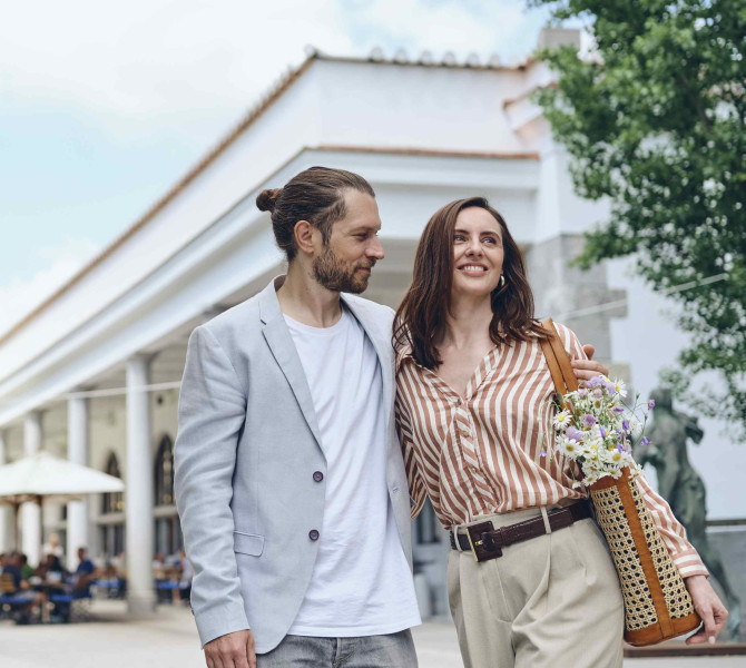 A man and a woman hug each other and walk past Plečnik's arcades. 