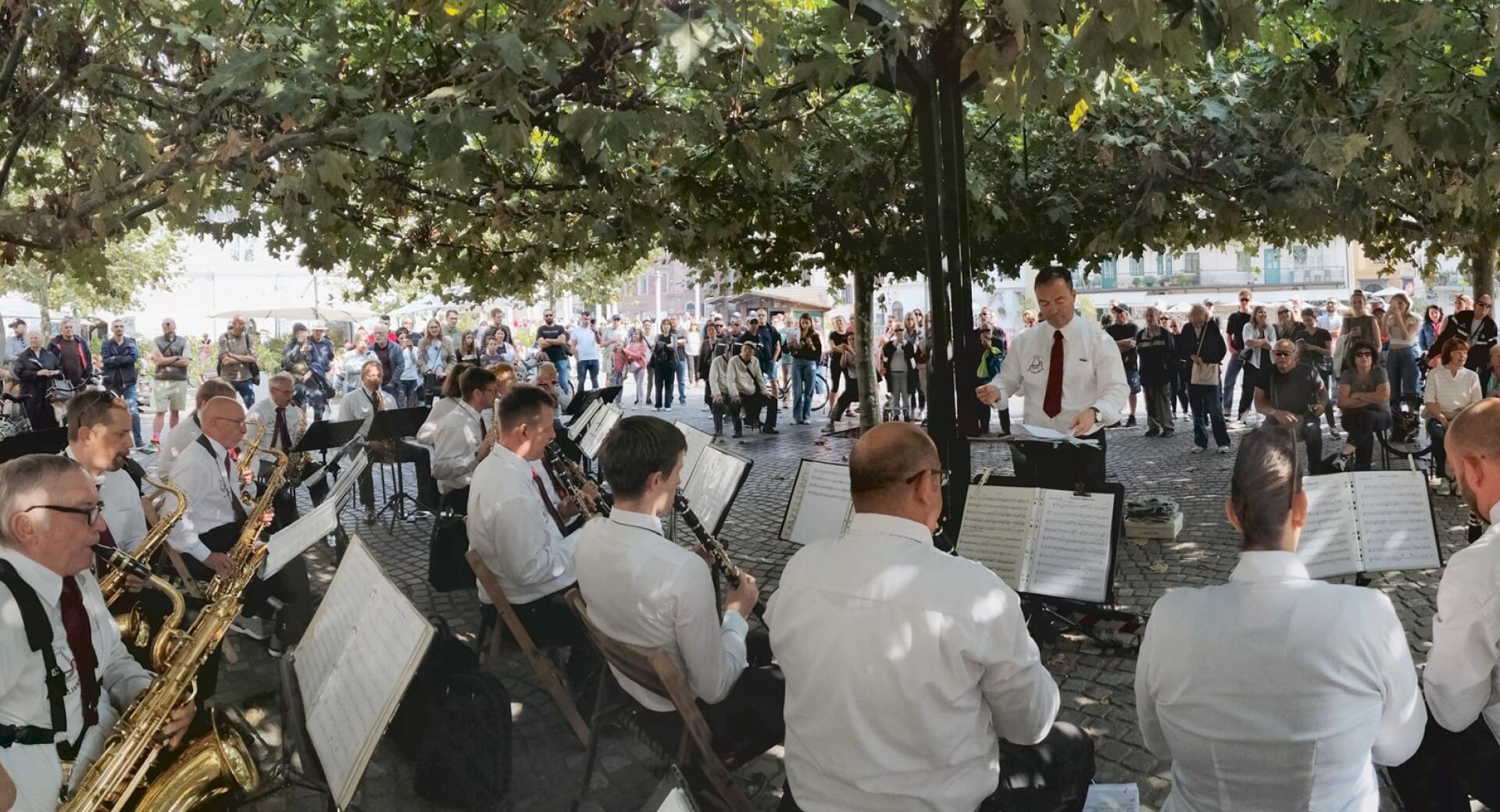 A wind band performs beneath leafy trees in a town square, entertaining a gathered crowd of spectators.
