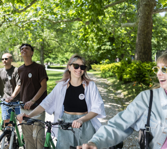 Four young people are walking with a bicycle along a path in a green park.