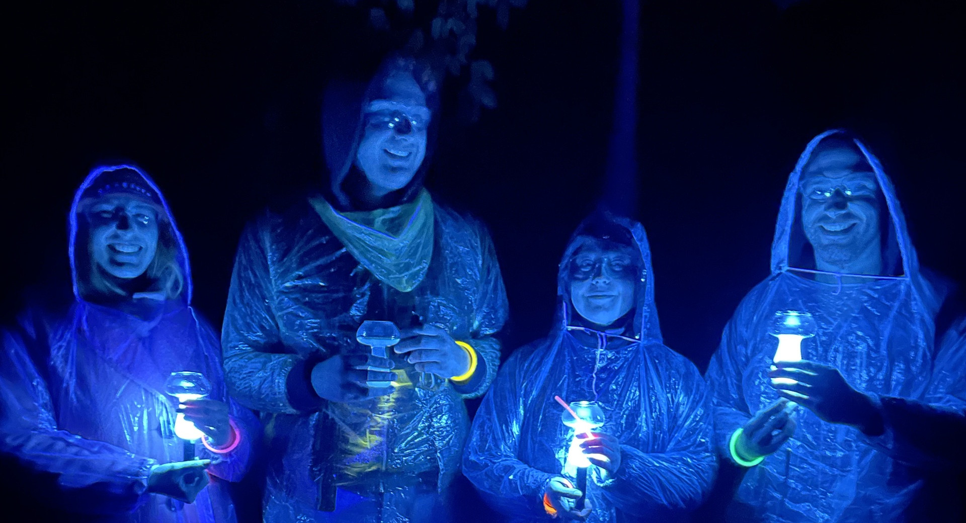 Four people in transparent rain ponchos stand in the dark, using UV lights to illuminate mushroom-shaped glasses in which the drink glows under UV light.