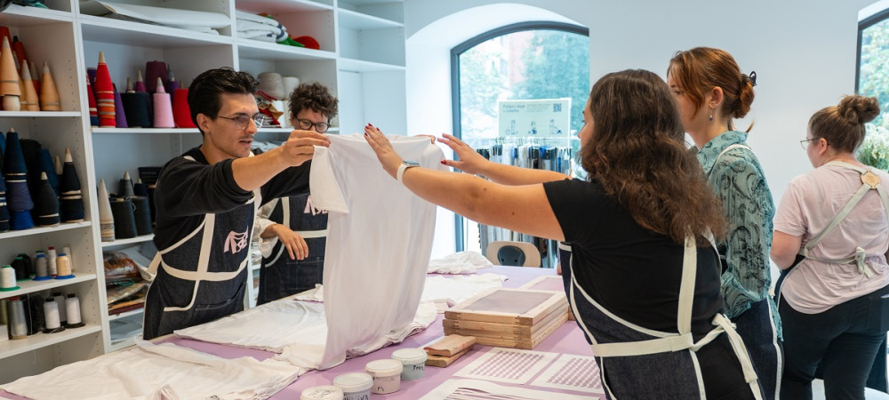 Un grande tavolo con colori e motivi per la stampa su magliette. Un uomo tiene una maglietta bianca sollevata mentre un gruppo di persone lo osserva.