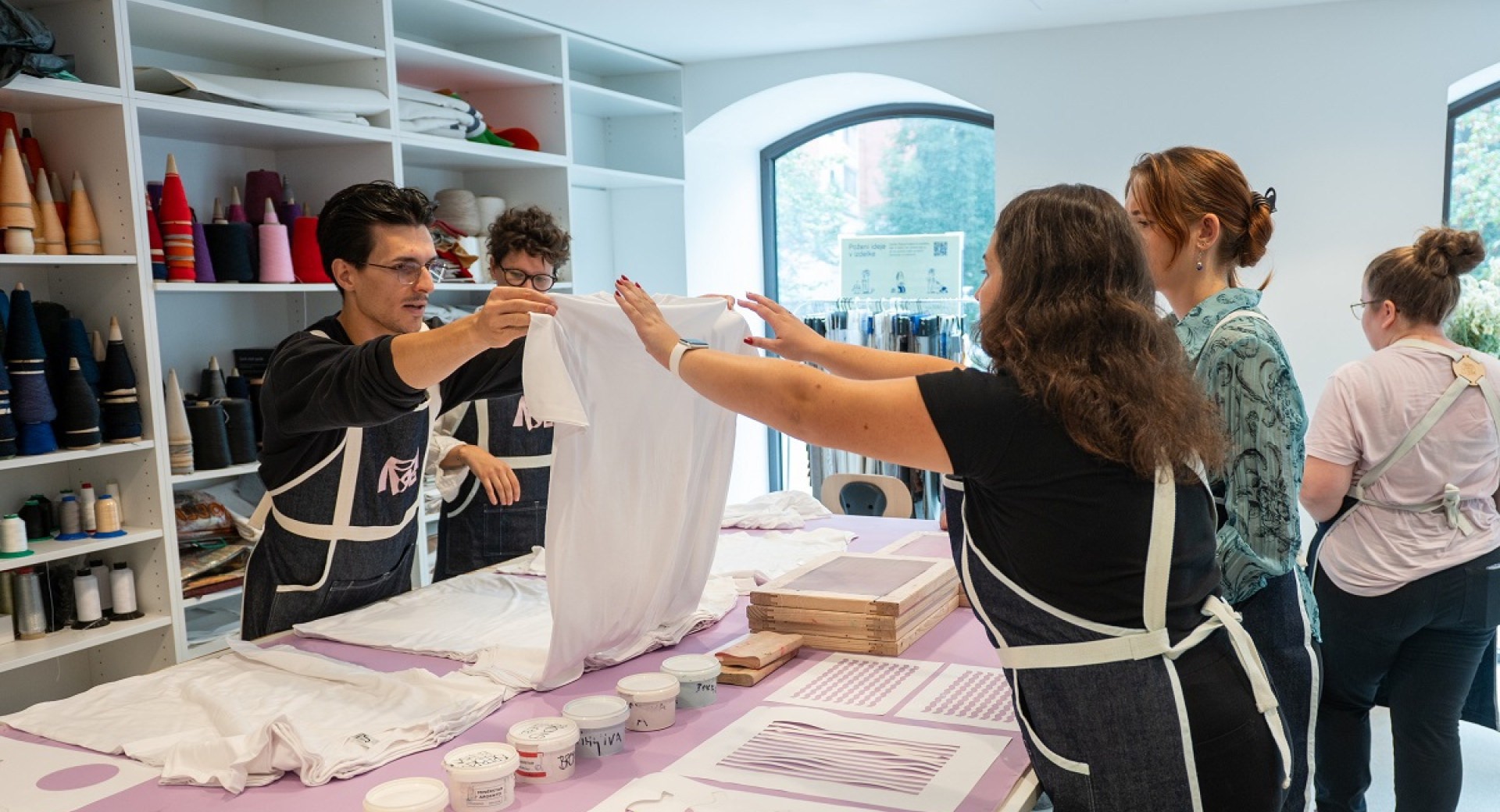 Un grande tavolo con colori e motivi per la stampa su magliette. Un uomo tiene una maglietta bianca sollevata mentre un gruppo di persone lo osserva.
