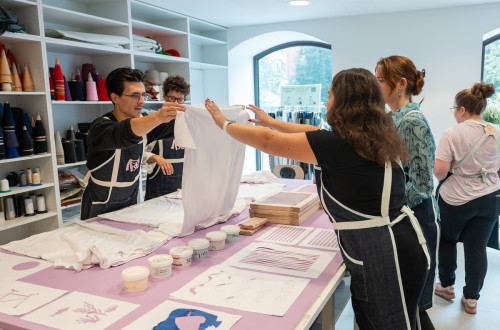 Velika miza z barvami in vzorci za tisk na majice. Moski drzi belo majico v zraku, skupina ljudi ga gleda.