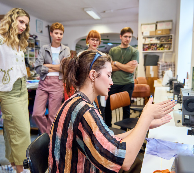 Four people are standing in a workshop while a woman is sitting and explaining something to them.