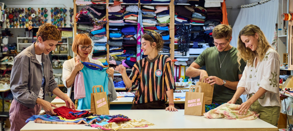Five people are standing in a room by a table with different types of fabric, getting ready to sew.