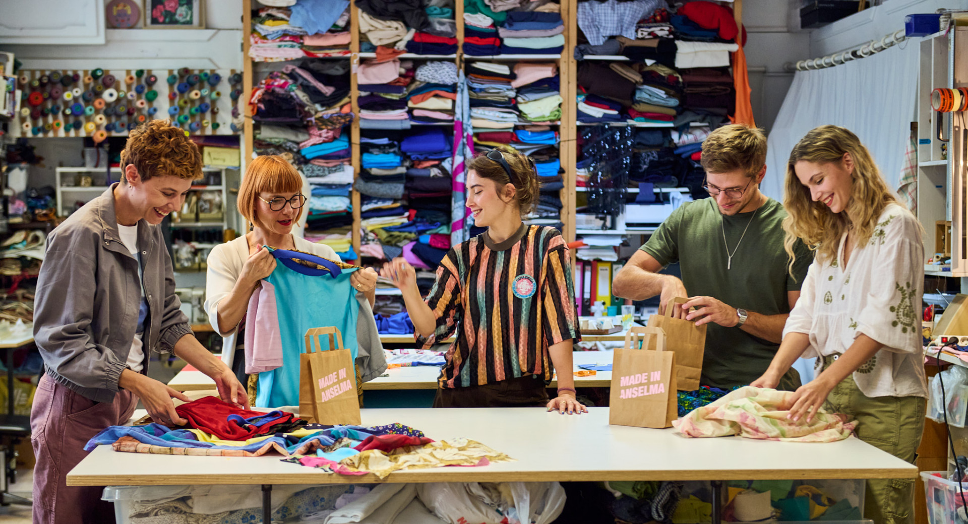 Five people are standing in a room by a table with different types of fabric, getting ready to sew.