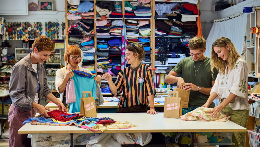 Five people are standing in a room by a table with different types of fabric, getting ready to sew.