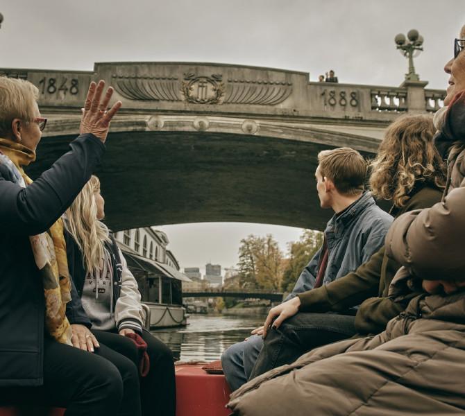 Ljudje sedijo na ladjici in opazujejo most, ki ga je ustvaril najbolj znani slovenski arhitekt Joze Plecnik. 