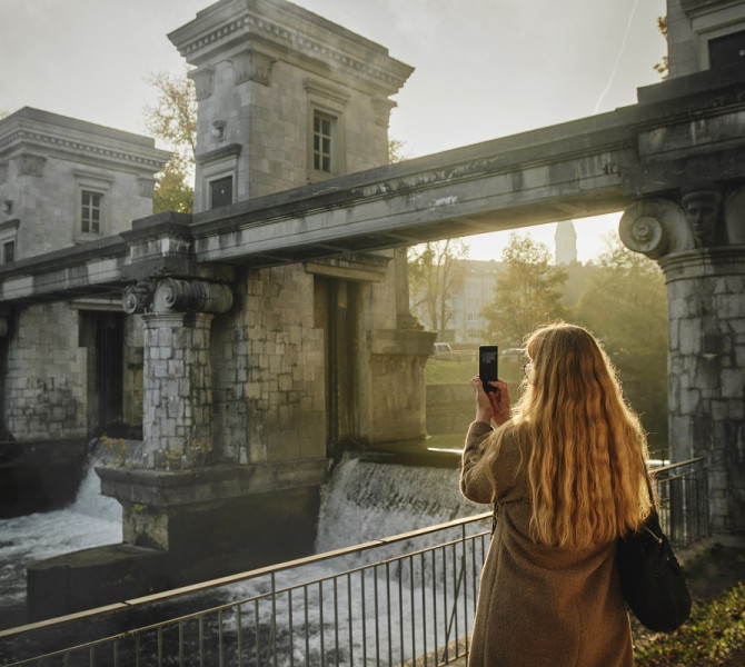 Zenska s svetlimi kodrastimi lasmi drzi v rokah mobilni telefon in fotografira zapornico na Ljubljanici.