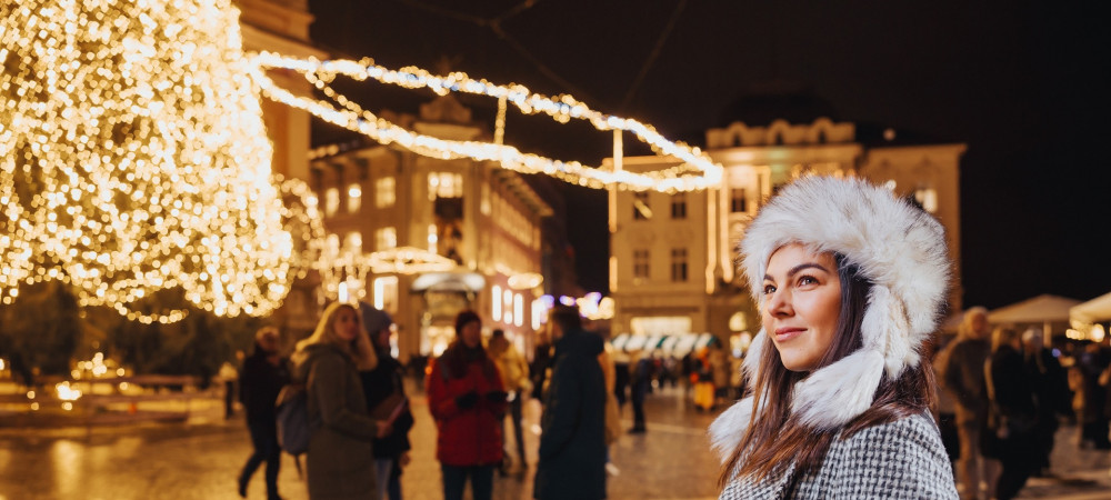 Eine Frau in einem grauen Mantel und einer weißen Pelzmütze steht inmitten einer festlich geschmückten Stadt.