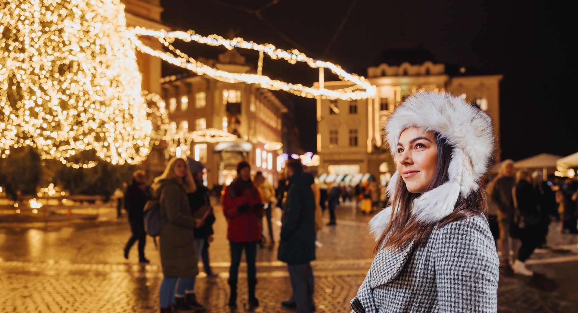 Zenska v sivem plascu z belo kucmo stoji sredi praznicno okrasenega mesta.