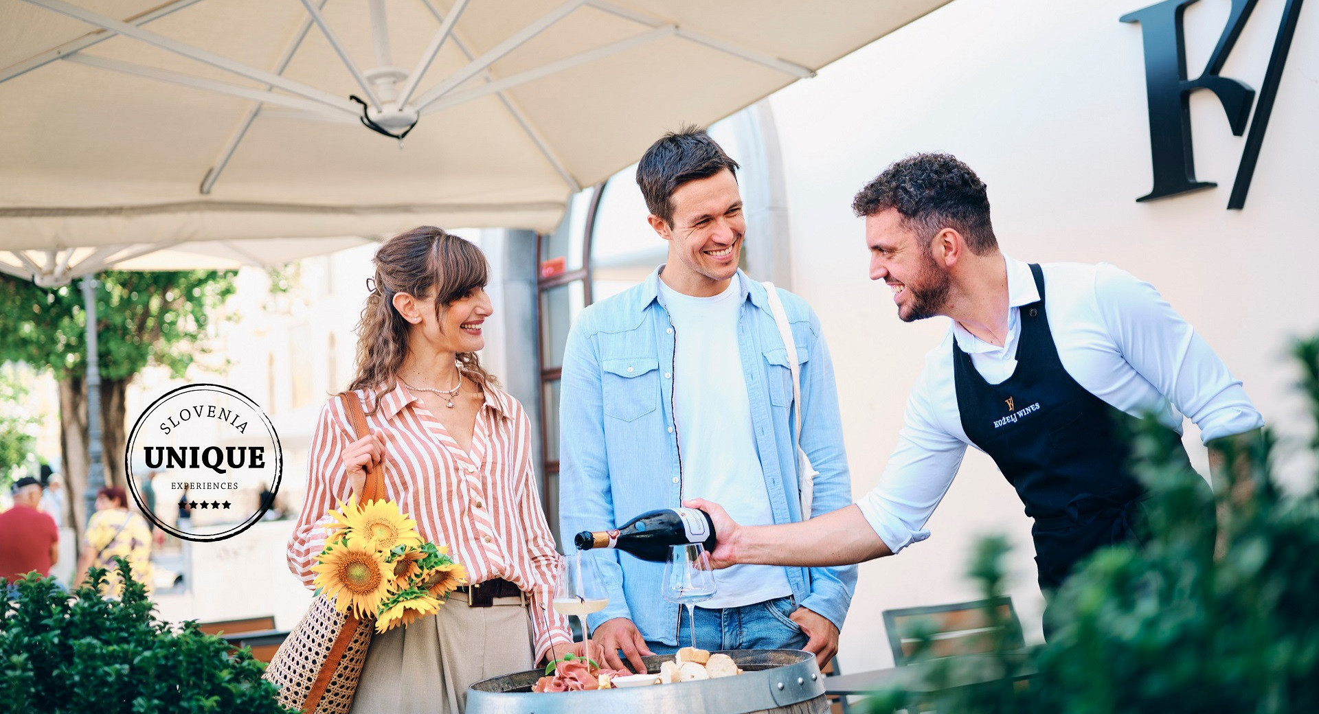 A smiling couple is tasting wine and food at the Ljubljana central market, accompanied by a sommelier presenting Kozelj wines.