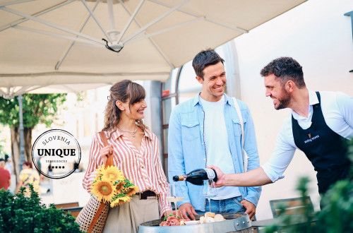 A smiling couple is tasting wine and food at the Ljubljana central market, accompanied by a sommelier presenting Kozelj wines.